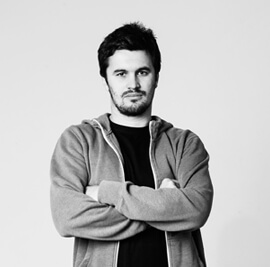 A black-and-white photo of a man with short, dark hair and a beard, standing with arms crossed. He is wearing a light jacket over a dark shirt and looking directly at the camera with a neutral expression. The background is plain and light-colored.