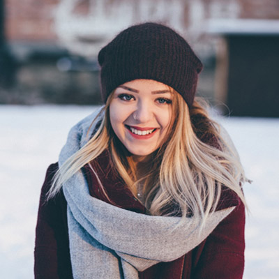 A woman with long blonde hair is smiling, standing outside in the snow. She is wearing a dark knit hat, a maroon coat, and a large gray scarf. The snowy background and her warm clothing suggest it's cold weather.