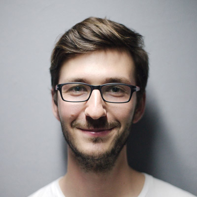 A man with short brown hair, a light beard, and black-rimmed glasses is smiling at the camera. He is wearing a white shirt, and the background is a neutral gray.