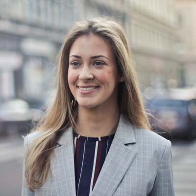 A woman with long, light brown hair is smiling while looking slightly to her right. She is wearing a light gray blazer over a striped shirt. The background is a blurred city street with some buildings and cars visible.