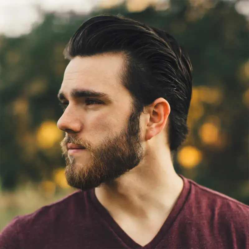 A person with dark, slicked-back hair, a full beard, and a mustache gazes to the left. They are wearing a maroon V-neck shirt, with window blinds in view behind them against the blurred outdoor setting of green and yellow hues.