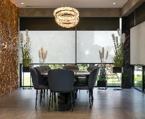 A modern dining room featuring a table with eight black chairs, a stylish chandelier, floor-to-ceiling windows with roller shades, and tall plants placed by the windows. The room has a neutral color palette and wooden floor with a nature-inspired accent wall on the left and sleek window blinds for added privacy.