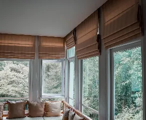 A cozy indoor seating area with large windows offering a view of lush greenery outside. The windows are adorned with beige window-blinds, and the bench below the windows is decorated with plush pillows, creating a serene and inviting atmosphere.