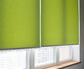 A close-up view showcases a set of green window blinds partially covering the windows of a room. The bottom part of the windows and the ledge are visible, letting in some natural light and revealing an urban landscape outside.