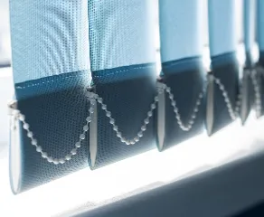 Close-up of window-blinds in shades of blue, with a white bead chain connecting the bottom of each slat. Bright light filters through the window, illuminating parts of the blinds and creating a soft glow.