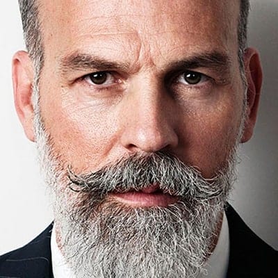 A close-up of a middle-aged man with a serious expression. He has a well-groomed gray beard and mustache, short hair, and is wearing a dark suit jacket with a white shirt, against a neutral background.