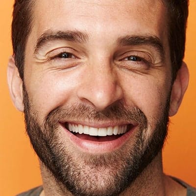 A man with a beard and short hair is smiling broadly against an orange background. He is wearing a grey shirt.