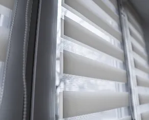 Close-up view of a modern window-blind setup with dual-layered, horizontal blinds partially opened, allowing light to filter through. The window frame appears to be white, and a chain for adjusting the blinds hangs on the left side.