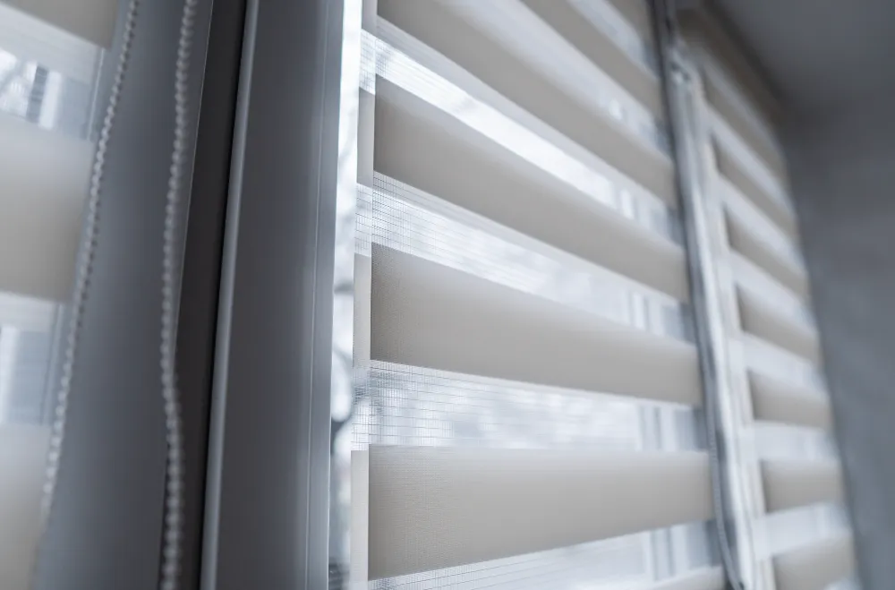 Close-up of modern window blinds with alternating translucent and opaque stripes, partially drawn to let in soft light. The blinds are in a room with a light-colored wall, creating a minimalist and contemporary feeling.