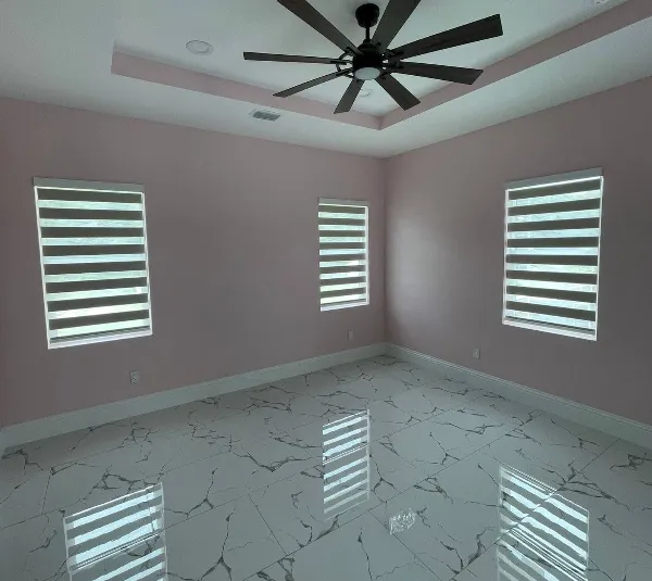 An empty room with pink walls, three windows adorned with stylish window-blinds, and a white marble floor accented by gray veins. The ceiling boasts a recessed design highlighted by a large black ceiling fan. Natural light gently filters in through the windows.