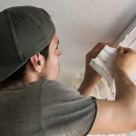 A person wearing a cap and gray t-shirt hangs curtains on a ceiling-mounted track, seamlessly complementing their expertise with window blinds installation. Standing on a ladder in the room's white walls, they carefully attach the pristine curtain to the railing.