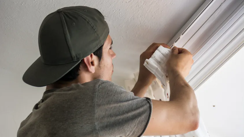 A person wearing a cap and gray t-shirt hangs curtains on a ceiling-mounted track, seamlessly complementing their expertise with window blinds installation. Standing on a ladder in the room's white walls, they carefully attach the pristine curtain to the railing.