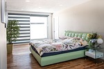 A modern bedroom with a green upholstered bed, floral comforter, and a white side table holding flowers and books. The room has wooden floors and a large window with striped blinds, allowing natural light in. A tall plant stands in the corner.