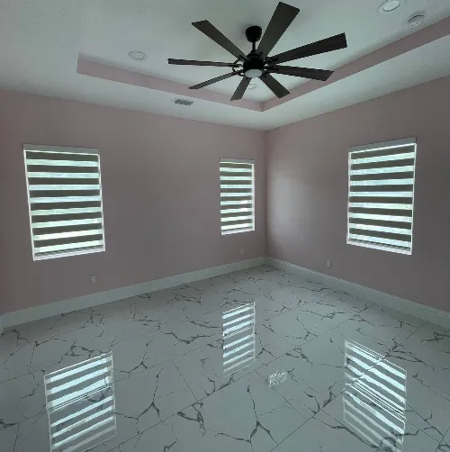 An empty room with glossy white marble floors and light pink walls showcases our exquisite window blinds. Three windows, adorned with striped blinds, allow soft light to filter in. A large black ceiling fan takes center stage above, adding a touch of elegance to the serene space.