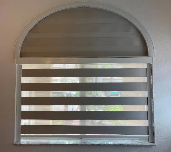 A window with horizontal striped window-blinds and an arched top section featuring matching shading. The blinds partially obscure the view outside, filtering light into the room.