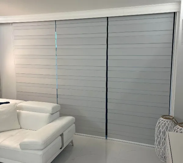 A modern living room featuring a white leather sectional sofa. The space boasts large sliding window blinds in grey, complementing the white walls and polished flooring. A decorative lantern sits gracefully on the floor beside the sofa.